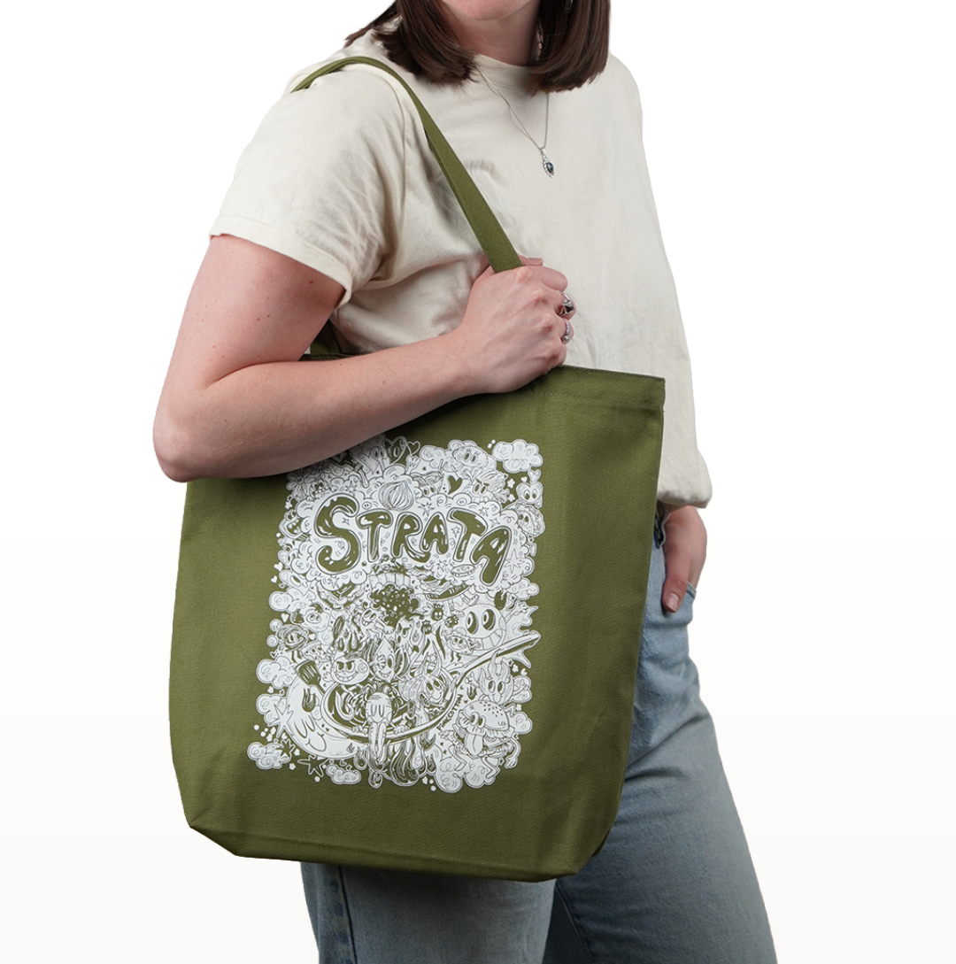 An olive green canvas tote bag with a white dreamy illustration and the word 'STRATA' printed on the front, carried by a model wearing a cream t-shirt and blue jeans against a white background.