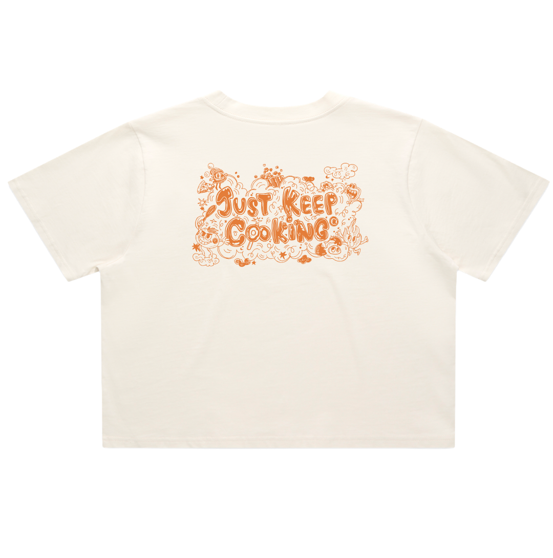 An off-white short-sleeve t-shirt with a retro-style orange graphic on the back reading 'JUST KEEP COOKING' surrounded by doodles of ingredients and utensils, shown against a plain white background.