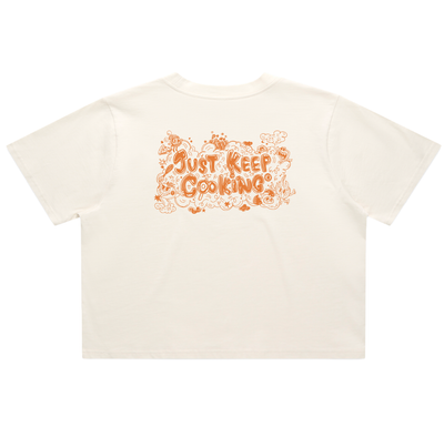 An off-white short-sleeve t-shirt with a retro-style orange graphic on the back reading 'JUST KEEP COOKING' surrounded by doodles of ingredients and utensils, shown against a plain white background.