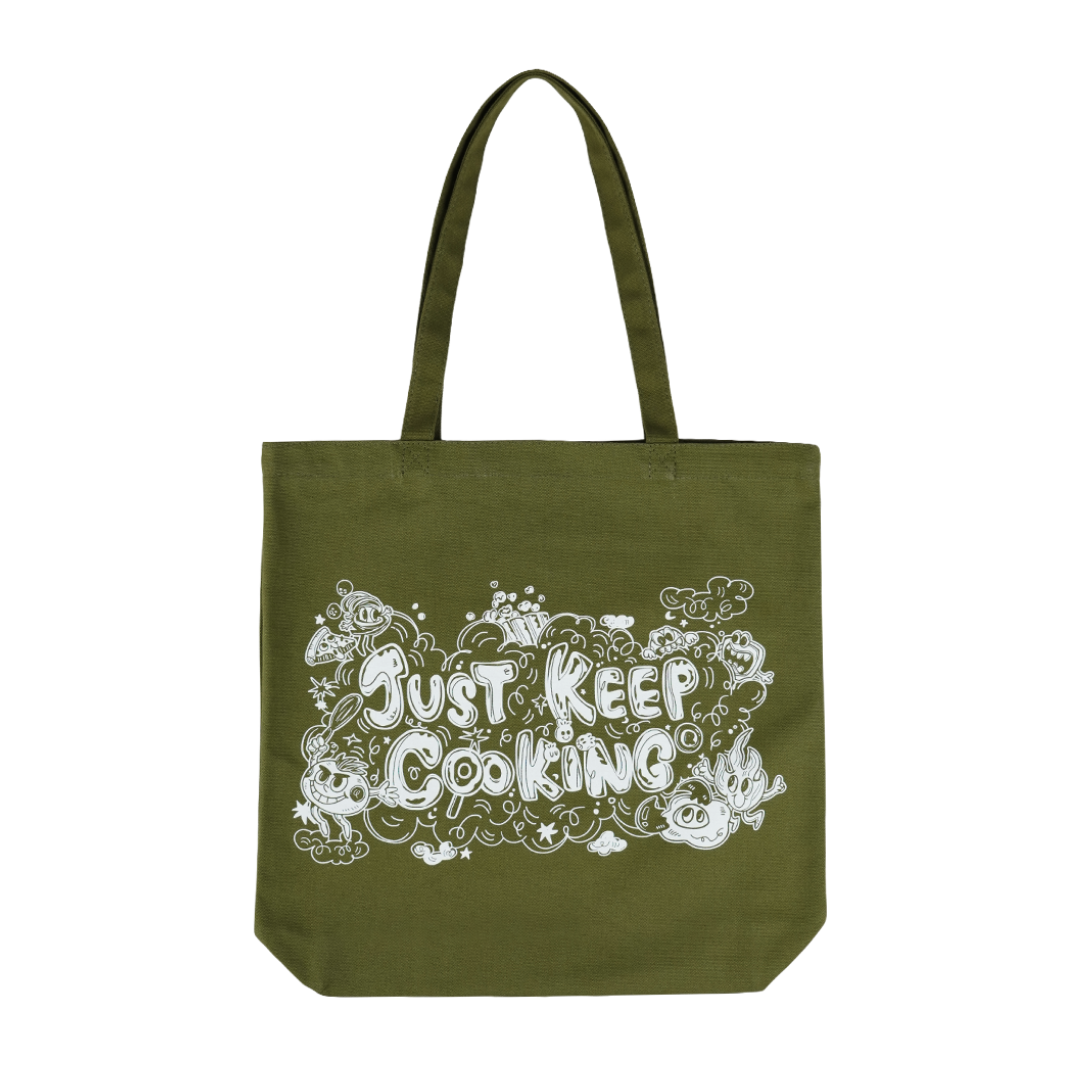 An olive green canvas tote bag with two handles, featuring a white graphic print on the front that reads 'Just Keep Cooking' surrounded by doodles of food and kitchen items, set against a plain white background.