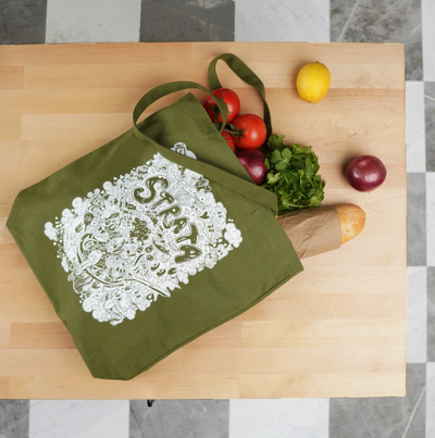 An olive green reusable tote bag with a white illustrated graphic and the word 'STRATA' printed on the front, shown on a light wood cutting board with groceries including tomatoes, parsley, a lemon, an apple, and a baguette.